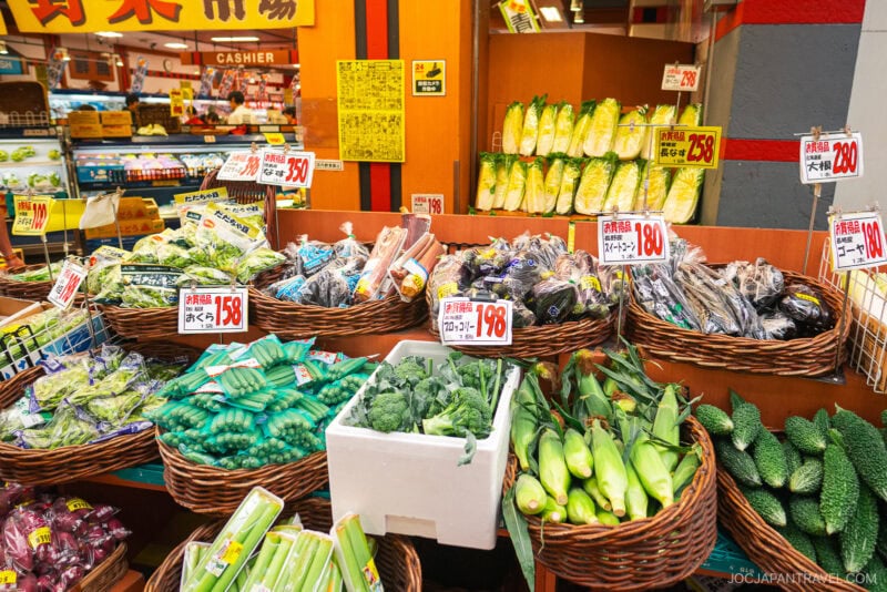 A colorful produce section at Kuromon Ichiba Market displays baskets of fresh vegetables, including corn, eggplants, cucumbers, and leafy greens, with handwritten price signs in Japanese yen.