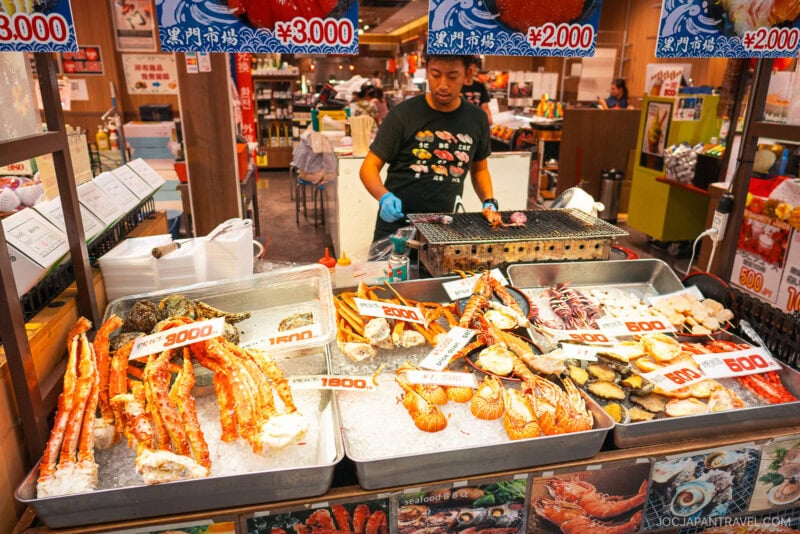 At Kuromon Ichiba Market, a seafood stall displays large crab legs, oysters, and scallops on ice with prices in yen. A person grills seafood behind the counter, surrounded by colorful signs and various market items.