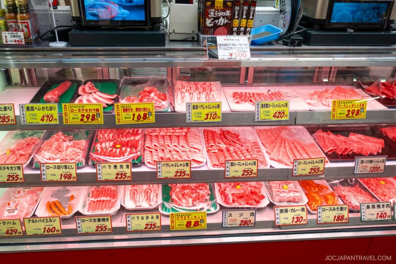 Glass display case filled with various cuts of raw meat, mostly beef and pork, each portion neatly arranged on trays with yellow price tags and Japanese labels in the brightly lit Kuromon Ichiba Market supermarket.