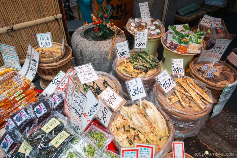 Various pickled vegetables and dried goods displayed in baskets and containers at a Japanese market stall in Kuromon Ichiba Market, with handwritten price signs in Japanese and vibrant packaging visible throughout.