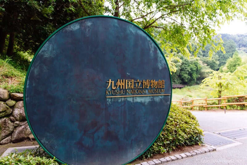 A large circular metal sign with gold lettering reads Kyushu National Museum in English and Japanese, set outdoors at the entrance to the Kyushu National Museum, surrounded by green trees and a scenic pathway.