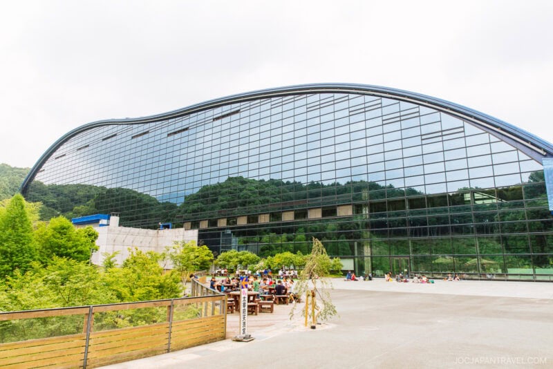 The large modern glass building with a wavy roof at the Kyushu National Museum reflects green trees and hills. In front, a group of people sit at outdoor tables on the spacious patio, surrounded by lush greenery in the bright scene.