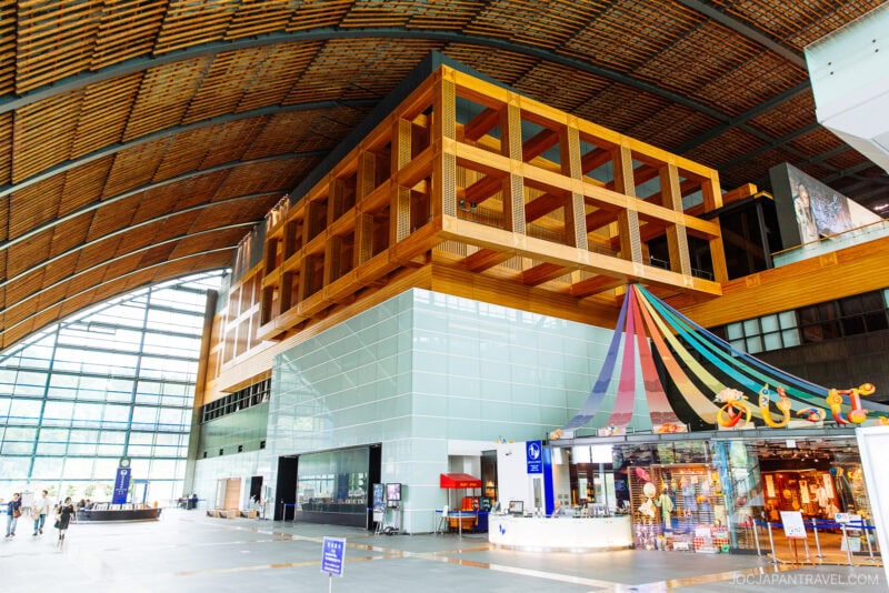 Spacious, modern interior at the Kyushu National Museum with a high arched ceiling, large windows, wood and glass accents, a colorful striped tent structure, and a small shop on the right. People are walking around the open area.