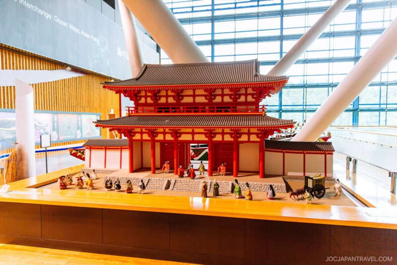 A detailed model of a traditional Japanese temple with red columns and a tiled roof, displayed indoors at the Kyushu National Museum, features small figurines arranged in front and is set against large windows and modern architectural elements.