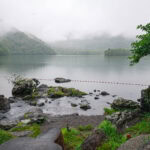 A misty Lake Chuzenji is surrounded by green forested hills, with rocks and a small tree in the foreground. A thin red-and-white rope stretches across the water, under an overcast sky with low-hanging clouds.