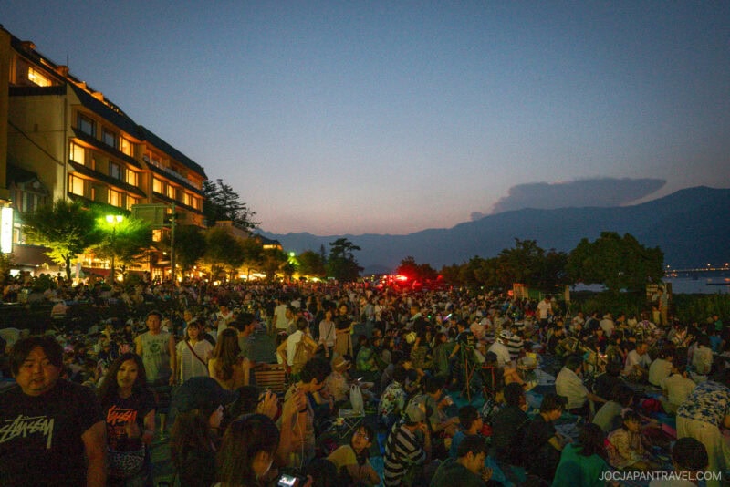 A large crowd gathers outdoors at dusk near a lit-up building, with mountains and a body of water visible in the background. The scene captures the vibrant atmosphere of the Lake Kawaguchi Fireworks Festival as evening approaches.
