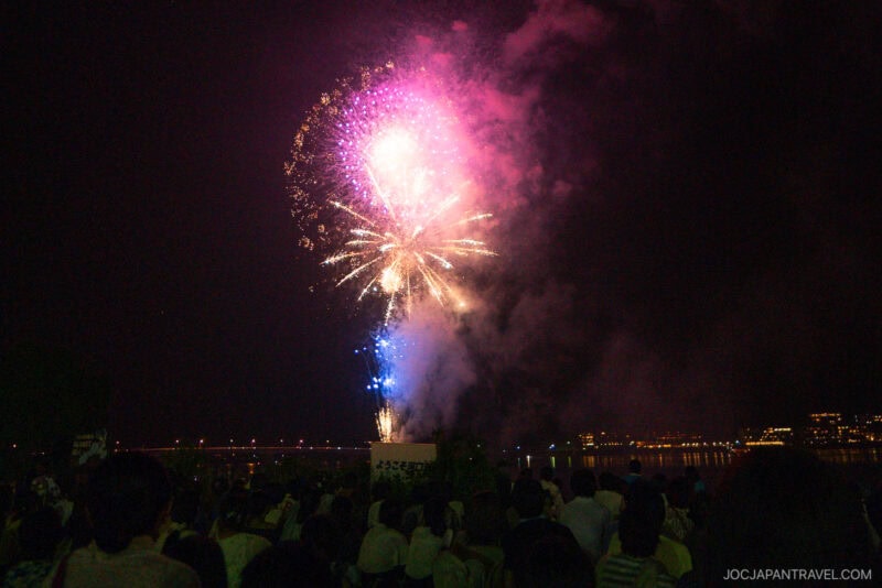 Colorful fireworks light up the night sky above a river at the Lake Kawaguchi Fireworks Festival, with a large crowd watching the display. City lights shimmer in the background, reflecting beautifully on the water.