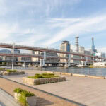 A wide riverside promenade in Meriken Park with benches and planters faces a waterway, elevated highways, and modern city buildings under a blue sky with wispy clouds.