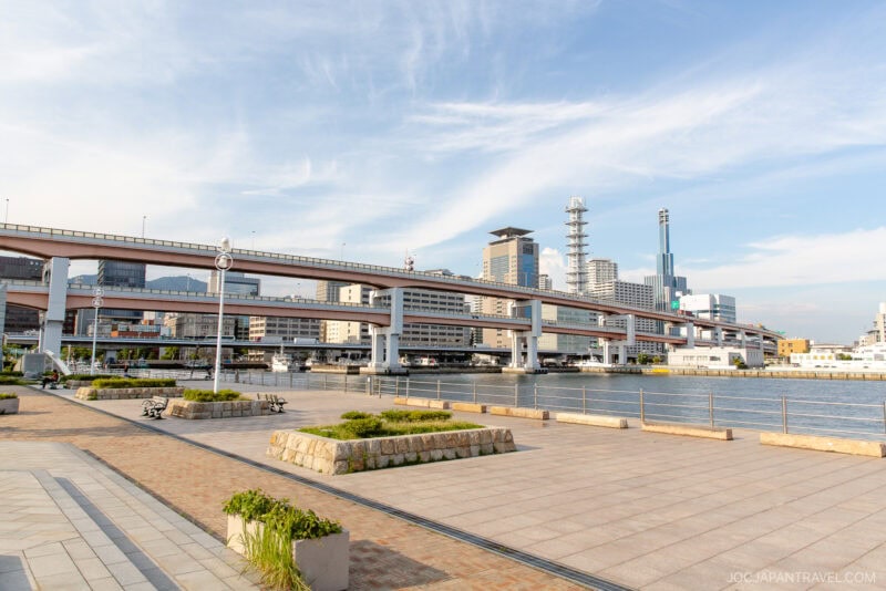 A wide riverside promenade in Meriken Park with benches and planters faces a waterway, elevated highways, and modern city buildings under a blue sky with wispy clouds.