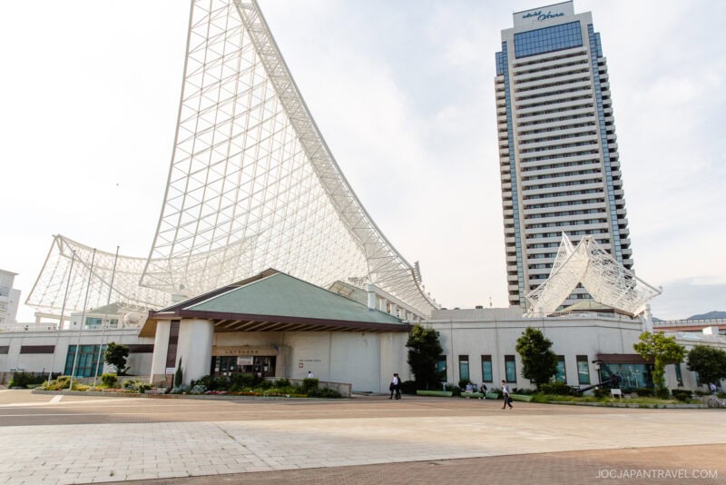 Modern building with a unique curved white steel structure in Meriken Park, beside a tall hotel tower; a few people walk in the open plaza area under a partly cloudy sky.