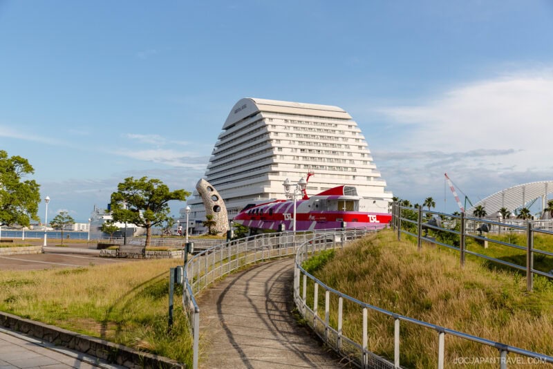 A curved path leads to a unique, white, pyramid-shaped building in Meriken Park. Nearby, a red sightseeing bus and a large fish sculpture stand out amid green grass, trees, and the blue sky surrounding the scene.