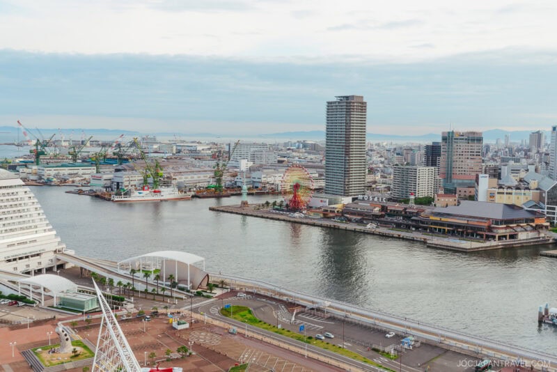 A cityscape view of Kobe, Japan, featuring modern buildings, Meriken Park, a ferris wheel, cranes, and a harbor waterfront under a cloudy sky. Roads and greenery are visible in the foreground.