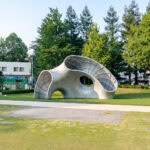 A modern, curved metallic sculpture sits on a grassy lawn in Midtown Garden, surrounded by trees, with a white building in the background and a paved walkway in front.