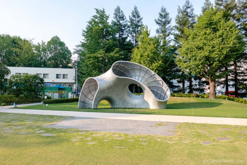 A modern, curved metallic sculpture sits on a grassy lawn in Midtown Garden, surrounded by trees, with a white building in the background and a paved walkway in front.