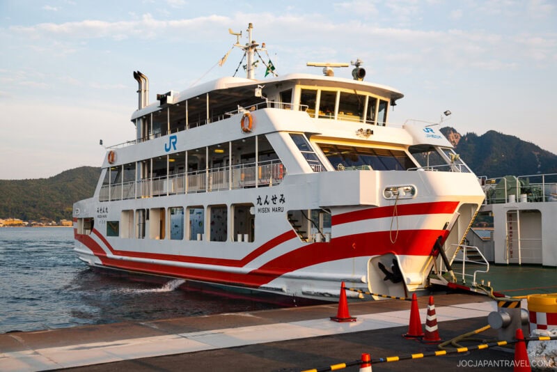 A white and red ferry named Miyajima Maru is docked at a pier in Miyajima, with mountains and water in the background. Orange traffic cones line the dock, and the ferry displays a JR logo.
