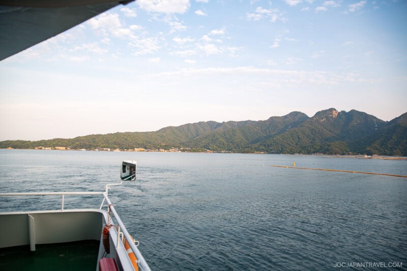 A scenic view from a boat deck looking toward Miyajimas mountainous coastline, under a partly cloudy sky, with calm blue water in the foreground.