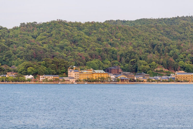 A scenic view of Miyajima&rsquo;s lakeside town, with traditional and modern buildings nestled at the base of a lush, green forested hill, seen across a calm body of water.