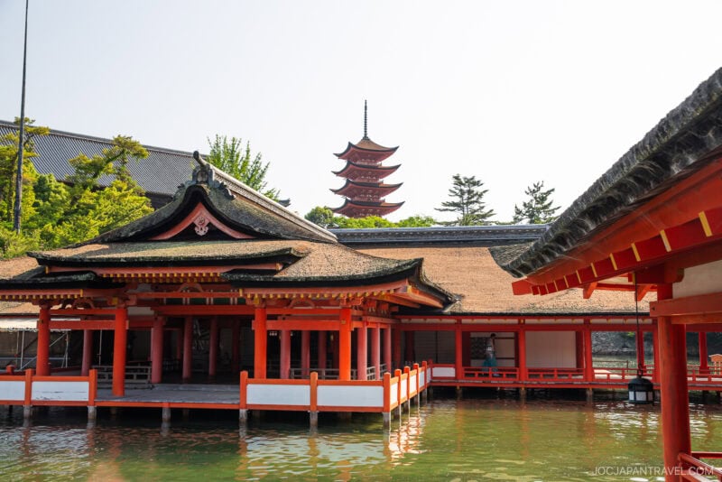 A traditional Japanese shrine with red pillars stands over water on Miyajima, with a multi-tiered pagoda and green trees visible in the background under a clear sky.