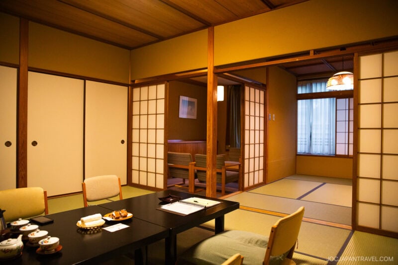 A traditional Japanese tatami room at Miyajima Grand Hotel Arimoto features sliding shoji doors, a low dining table with tea and snacks, and a seating area by the window. Warm lighting creates a cozy atmosphere.