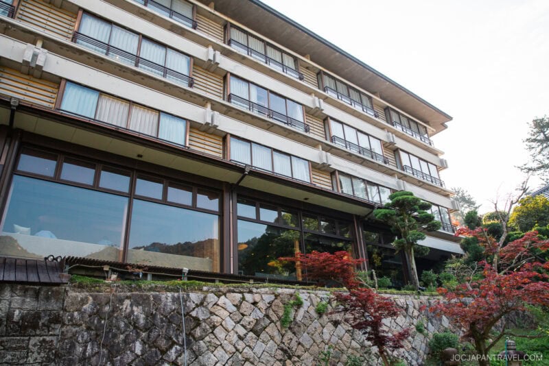 A multi-story building with large windows, wooden and white exterior walls, and a stone retaining wall, Miyajima Grand Hotel Arimoto is surrounded by green trees and red-leaved plants.