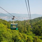 A green cable car travels above the lush forested hills of Mount Misen, with a distant city and mountains visible across a body of water under a clear blue sky.