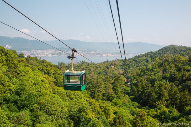 A green cable car travels above the lush forested hills of Mount Misen, with a distant city and mountains visible across a body of water under a clear blue sky.