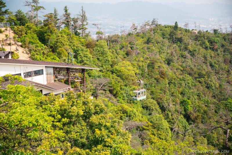 A cable car travels over the lush green forested hills of Mount Misen near a station building, with mountains and a hazy cityscape visible in the background.