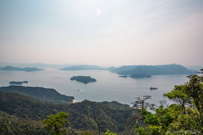 View of a calm, expansive bay with scattered small islands, surrounded by forested hills under a hazy sky; Mount Misen rises in the distance, a ship is visible on the water, and greenery fills the foreground.