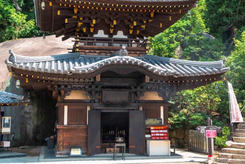 A traditional Japanese temple with ornate wooden architecture and a curved tiled roof sits at the base of Mount Misen, surrounded by greenery and large rocks. Candles glow inside, while signs are placed outside the entrance on a sunny day.