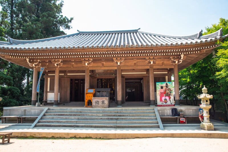 Traditional Japanese temple with a tiled roof, wooden pillars, and steps leading to the entrance. A lantern and colorful poster stand by the entrance, surrounded by greenery and trees beneath the clear sky of Mount Misen.