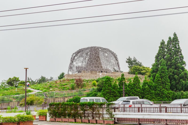 A large hexagonal metal dome structure sits on a hillside of Mount Rokko, surrounded by greenery and trees, with parked cars and a fence in the foreground on an overcast day.