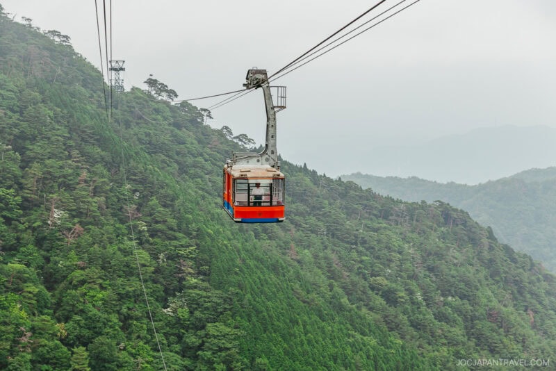 A red and white cable car travels over the lush, green forested slopes of Mount Rokko on a cloudy day.