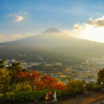 Mt. Fuji Panoramic Ropeway (富士山パノラマロープウェイ) – Scenic Views from Mt. Tenjo Mount Fuji rises above a town and forest with autumn foliage at sunset. People stand at a scenic overlook near the Mt. Fuji Panoramic Ropeway, surrounded by colorful statues among the trees under a partly cloudy sky.
