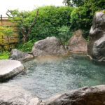 Musouen Hotel (山のホテル 夢想園) – A Serene Ryokan Stay in Yufuin An outdoor Japanese onsen at Musouen Hotel, surrounded by large rocks, green bushes, and a bamboo fence. Steam rises gently from the clear hot spring water, with a wooden bucket resting at the pools edge.