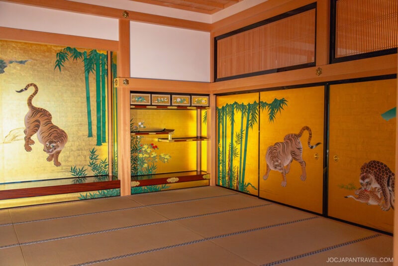 A traditional Japanese room with tatami mats and sliding doors, decorated with tiger and bamboo artwork on a gold background, illuminated by natural light—reminiscent of the elegant interiors found in Nagoya Castle.