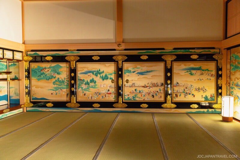 A traditional Japanese room with tatami mats and ornate wall panels depicting detailed historical scenes of Nagoya Castle in gold and vivid colors, illuminated by soft warm lighting.