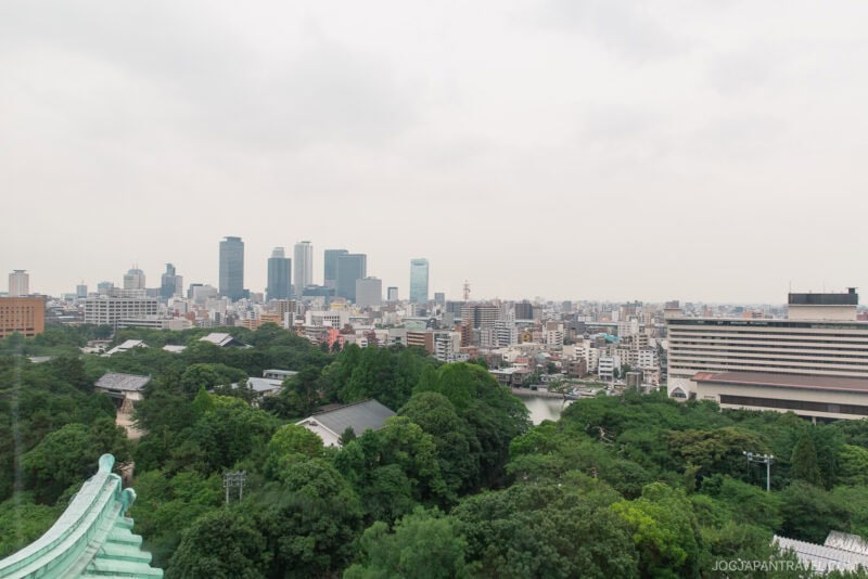 A cityscape view of Nagoya, Japan, featuring modern skyscrapers, mid-rise buildings, lush green trees, and part of Nagoya Castle&rsquo;s traditional-style roof in the foreground under a cloudy sky.