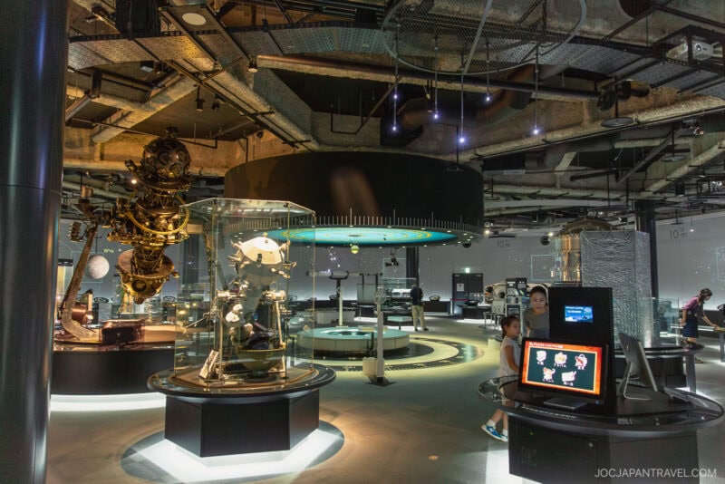 A modern science museum exhibit at the Nagoya City Science Museum features interactive displays, a large mechanical device, and information screens. Visitors explore industrial-style ceilings and dramatic lighting throughout the space.
