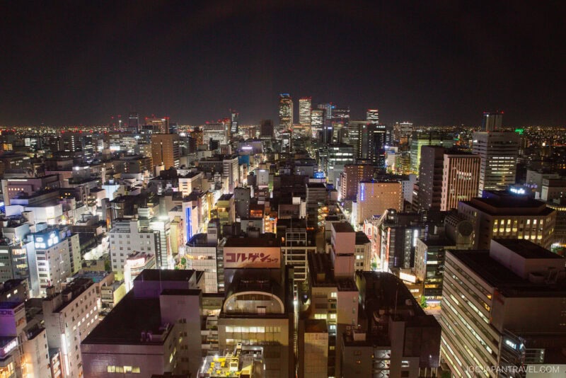 Aerial view of a brightly lit cityscape at night, featuring Nagoya TV Tower among numerous tall buildings, illuminated signs, and distant skyscrapers under a dark sky.
