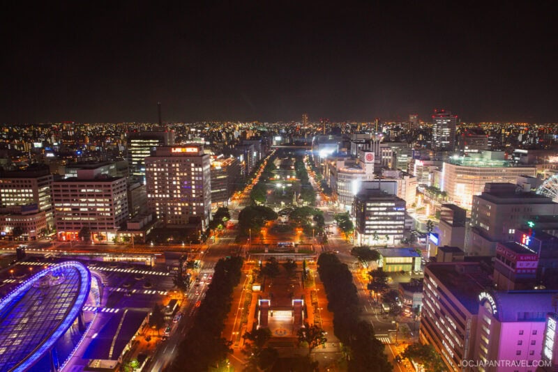 A vibrant cityscape at night with brightly lit buildings, tree-lined streets, and the Nagoya TV Tower glowing purple on the left, all set against a dark sky filled with city lights in the distance.