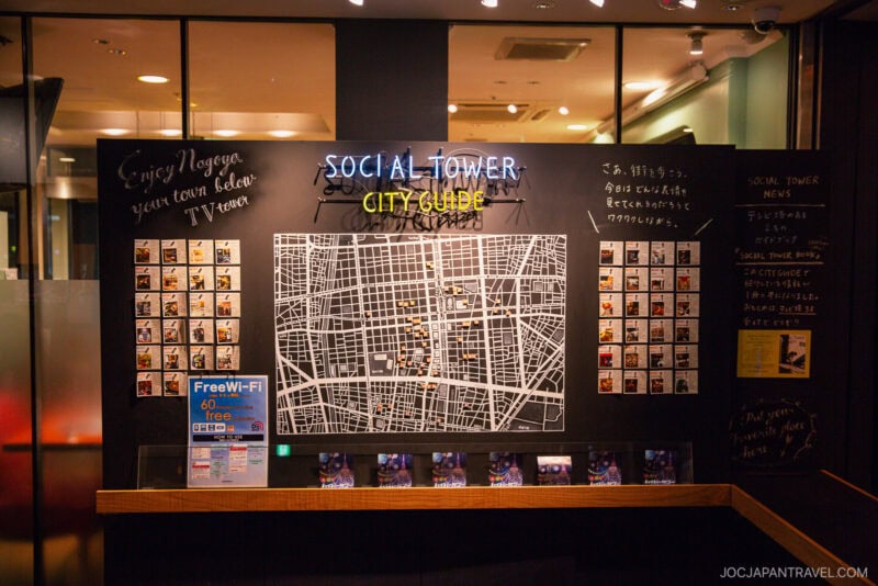 A brightly lit display board titled SOCIAL TOWER CITY CLUB, located near Nagoya TV Tower, features a city map, polaroid photos, handwritten notes in Japanese, and a sign advertising free Wi-Fi inside a modern building.