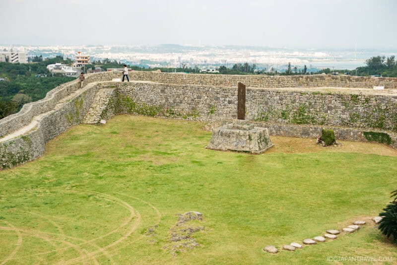 A wide view of Nakagusuku Castles historic stone wall enclosing a grassy area with a stone monument in the center; city buildings and the ocean are visible in the distance.
