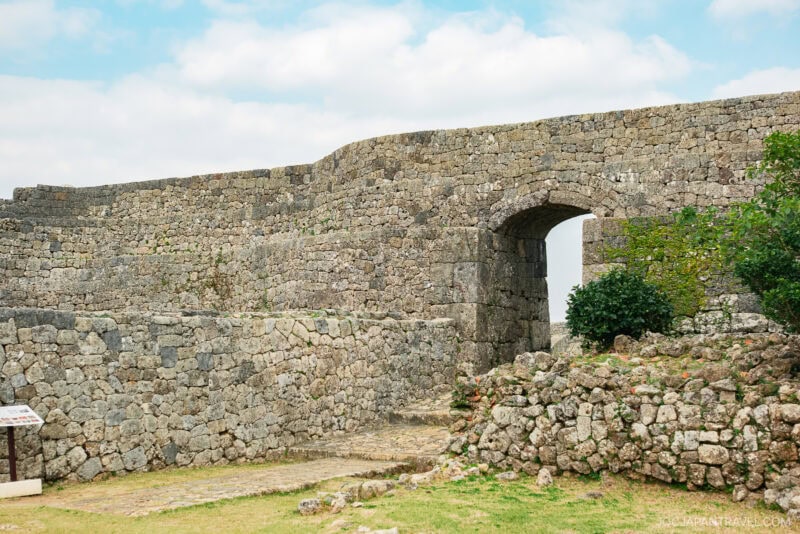 An ancient stone wall and arched gateway of Nakagusuku Castle stand under a partly cloudy sky, with patches of grass and small bushes growing nearby.