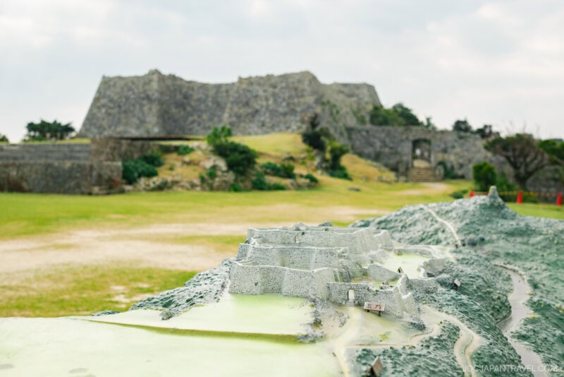A close-up of a scale model of Nakagusuku Castle is shown outdoors, with the actual historic castle ruins blurred in the background on a grassy landscape under a cloudy sky.