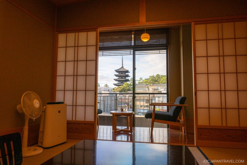 A traditional Japanese room at Nara Asukasou Ryokan features shoji doors open to a balcony with views of a pagoda and trees. The room includes a table, chair, fan, and air purifier, all bathed in gentle natural light.