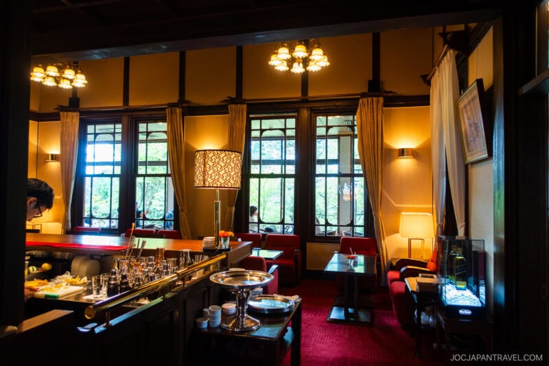Elegant lounge with large windows, red carpet, and classic wooden furniture at Nara Hotel. A person stands at a bar with various drinks. Warm lighting and lush greenery are visible outside the windows.