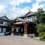 Nara Hotel and Ryokan Experiences A traditional Japanese building with ornate wooden architecture and tiled roofs, surrounded by greenery beneath a cloudy sky, with a few people walking nearby—reminiscent of the historic Nara Hotel.