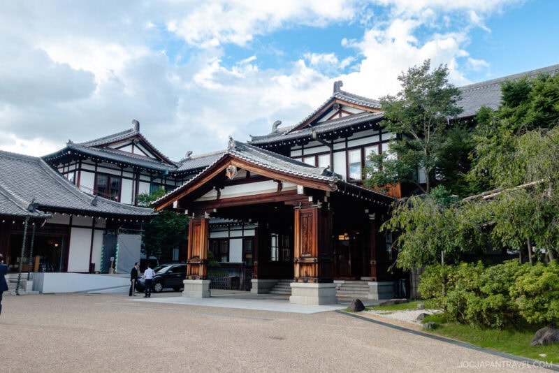 A traditional Japanese building with ornate wooden architecture and tiled roofs, surrounded by greenery beneath a cloudy sky, with a few people walking nearby—reminiscent of the historic Nara Hotel.