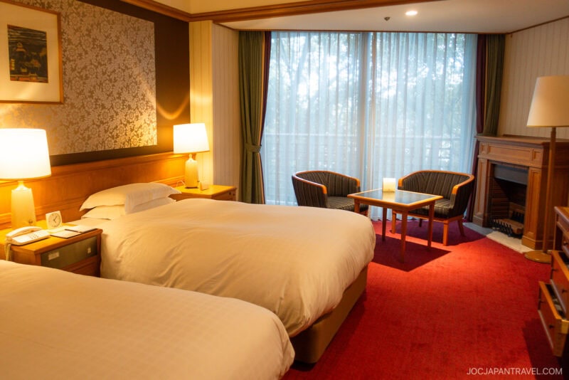 Hotel room at Nara Hotel with two double beds, a bedside table with lamps, a small sitting area by large windows with sheer curtains, and a red carpeted floor.