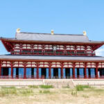 Nara Palace Site (Heijo Palace / 平城宮跡) A large, traditional Japanese building at the Nara Palace Site with red columns, white walls, and a dark tiled roof stands against a clear blue sky, with grass in the foreground.